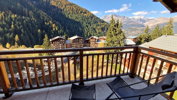 Un balcon avec deux fauteuils de jardin, donnant sur une région montagneuse parsemée de bâtiments.