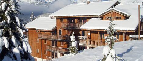 A wooden chalet with multiple balconies and a snow-covered roof, surrounded by snow and mountains.