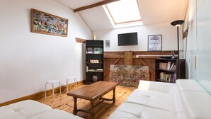 Un salon de style industriel, avec un plafond en pente, une table en bois, des canapés blancs, un téléviseur fixé au mur et une bibliothèque.