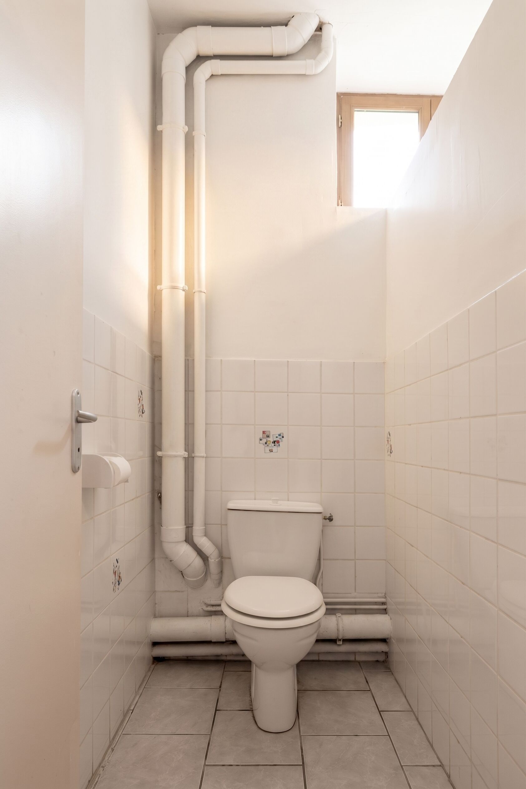 Een kleine badkamer met toilet, witte betegelde wanden en een zichtbaar leidingwerk.