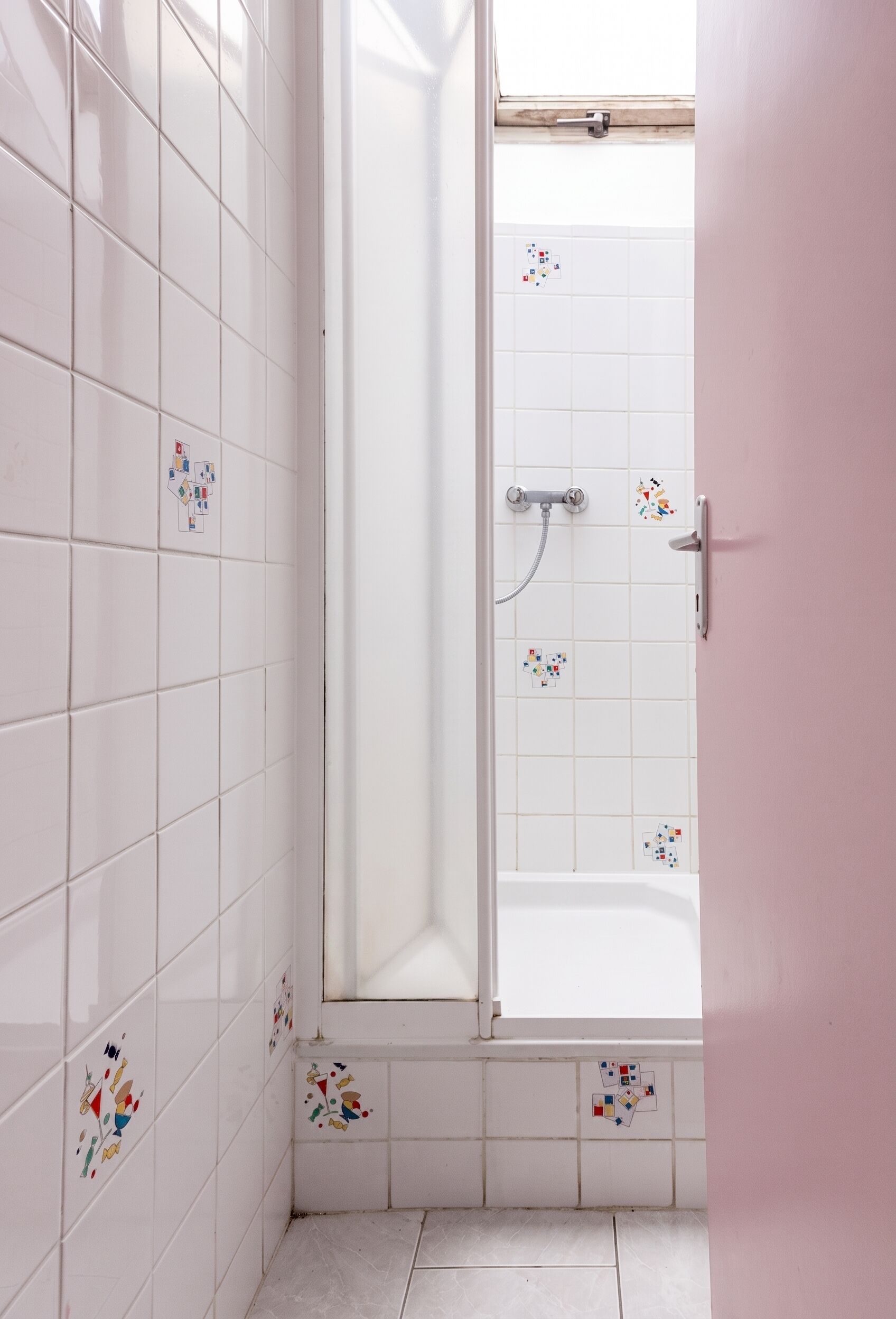 Ett badrum med vita kakelväggar, en dusch med en glasdörr och en rosa dörr.