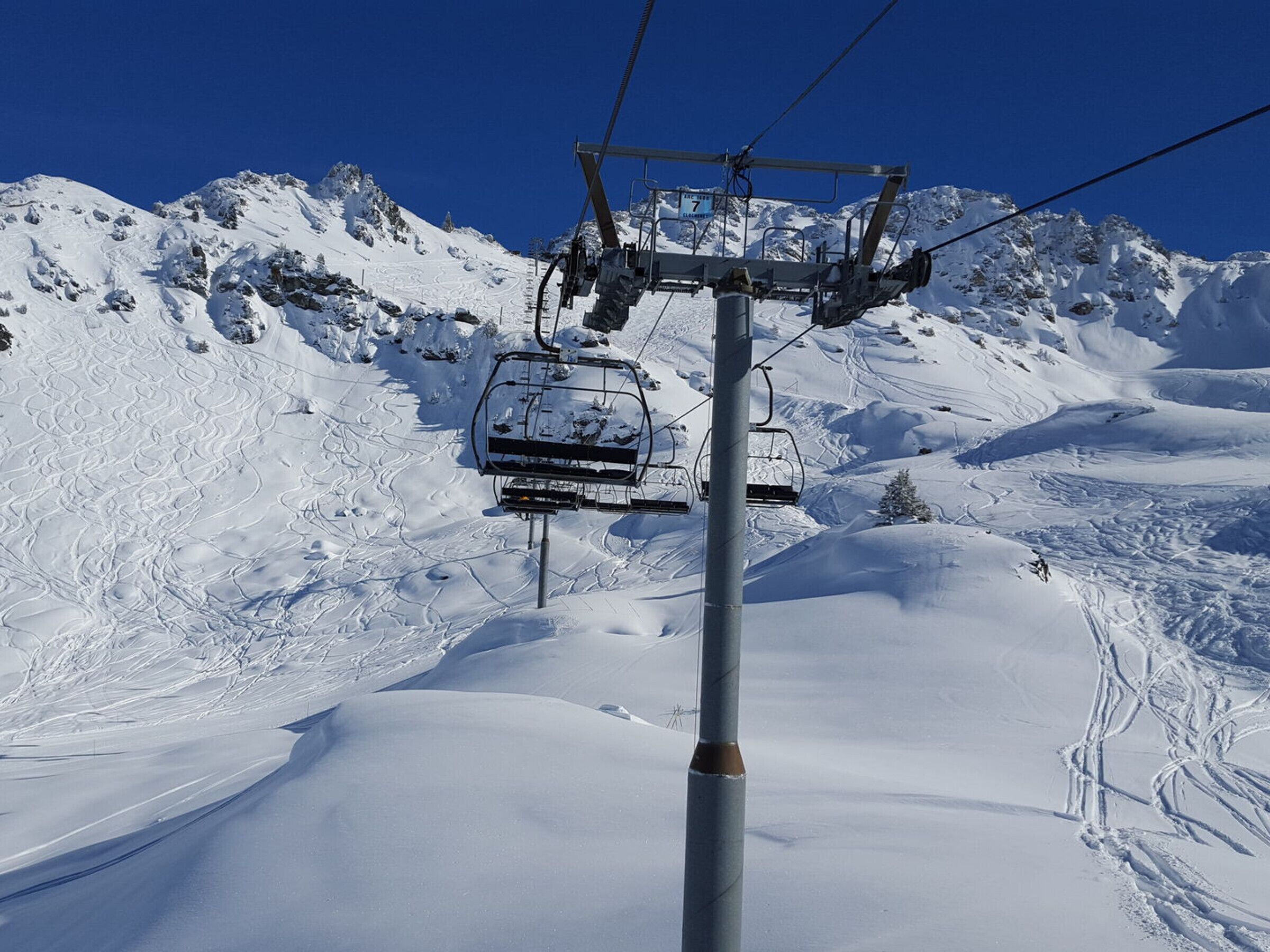 A ski lift chair suspended from a cable system, with a snowy mountain landscape in the background.