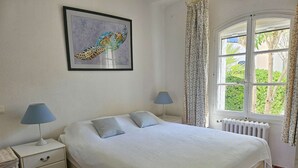 Une chambre à coucher avec un lit, deux tables de chevet, une commode, un radiateur, une fenêtre avec des rideaux et un tableau encadré au mur.