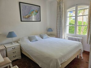 A bedroom with a bed, two bedside tables, a nightstand, a radiator, a window with curtains, and a framed picture on the wall.