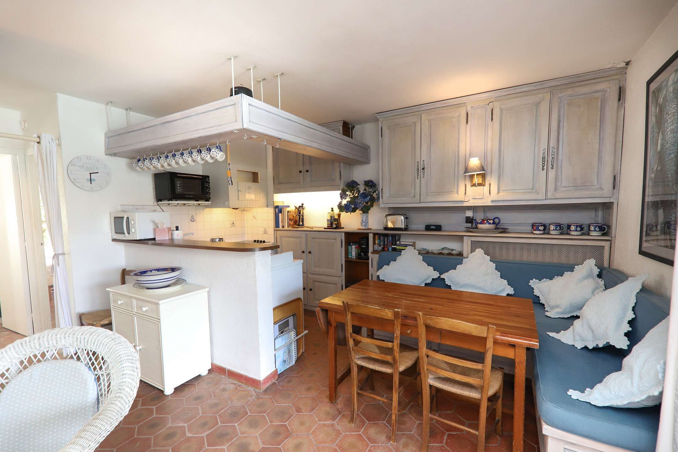 Une cuisine compacte avec un coin repas, des meubles blancs, un four à micro-ondes et un banc bleu avec un coussin.