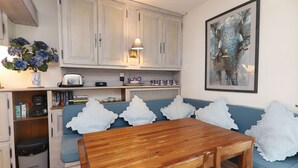 Une cuisine compacte avec des meubles blancs, une table à manger en bois, un canapé bleu agrémenté de coussins blancs et, au mur, un tableau représentant un éléphant.