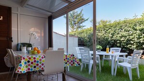 An open patio with a dining table and chairs, a small table with fruits and a flower, and a view of a garden and trees. - Beachfront Villa with Garden & Pool, 200m from Seignosse Beach (Seignosse)