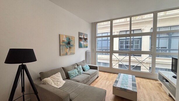 Ein Wohnzimmer mit grauem Sofa, einer schwarzen Stehlampe und einem Couchtisch. An der Wand hängen zwei abstrakte Gemälde.