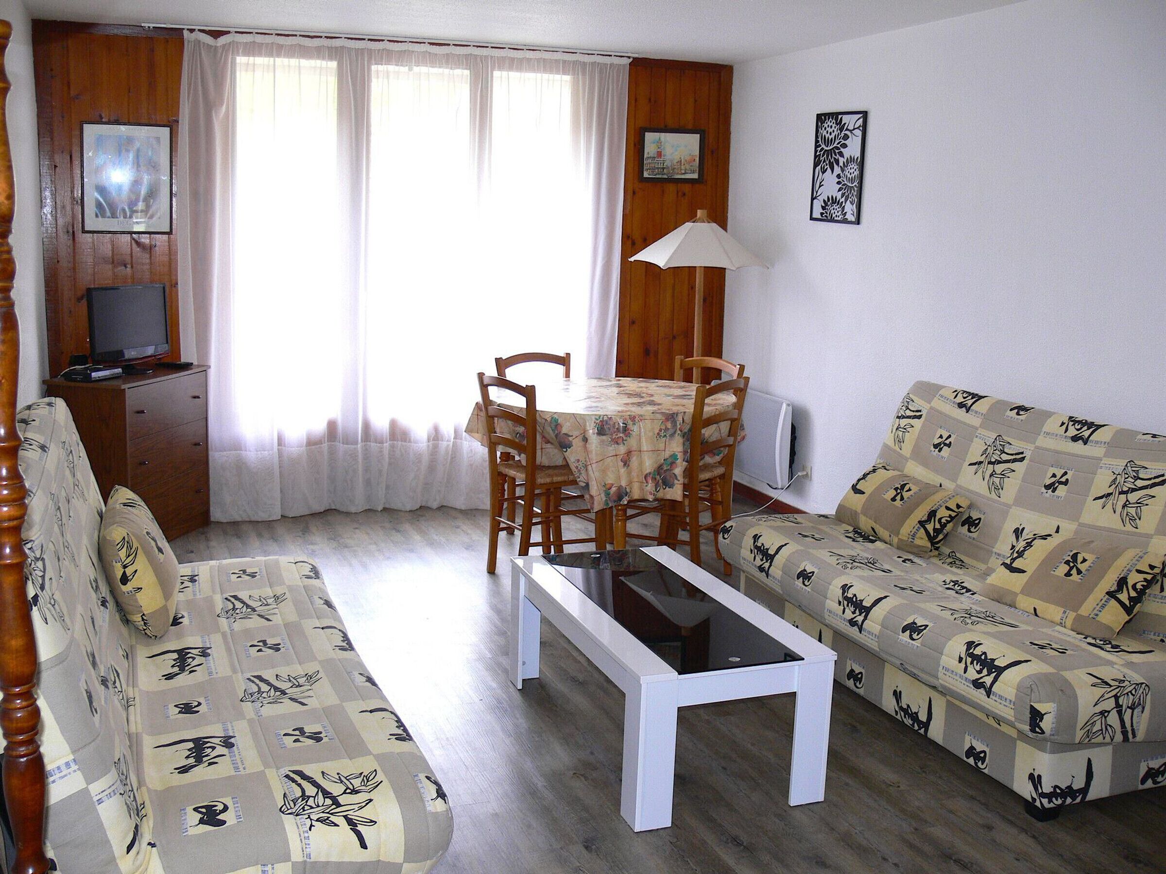 Ein Wohnzimmer mit einer Couch, einem Esstisch mit Stühlen, einem Couchtisch, einem Fernseher auf einer Holzkommode und einem Fenster mit Vorhängen.