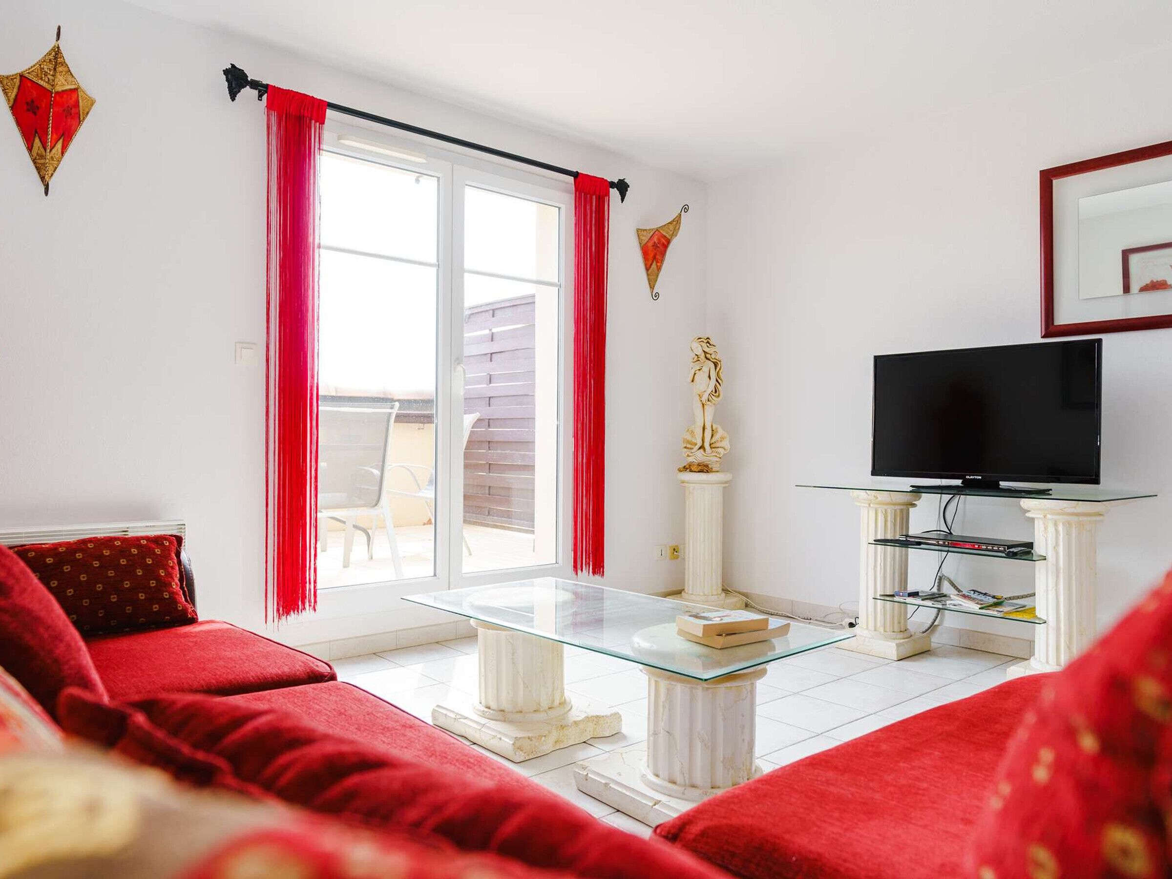 A living room with a glass coffee table, a sofa, a TV on a stand, and a balcony with red curtains.