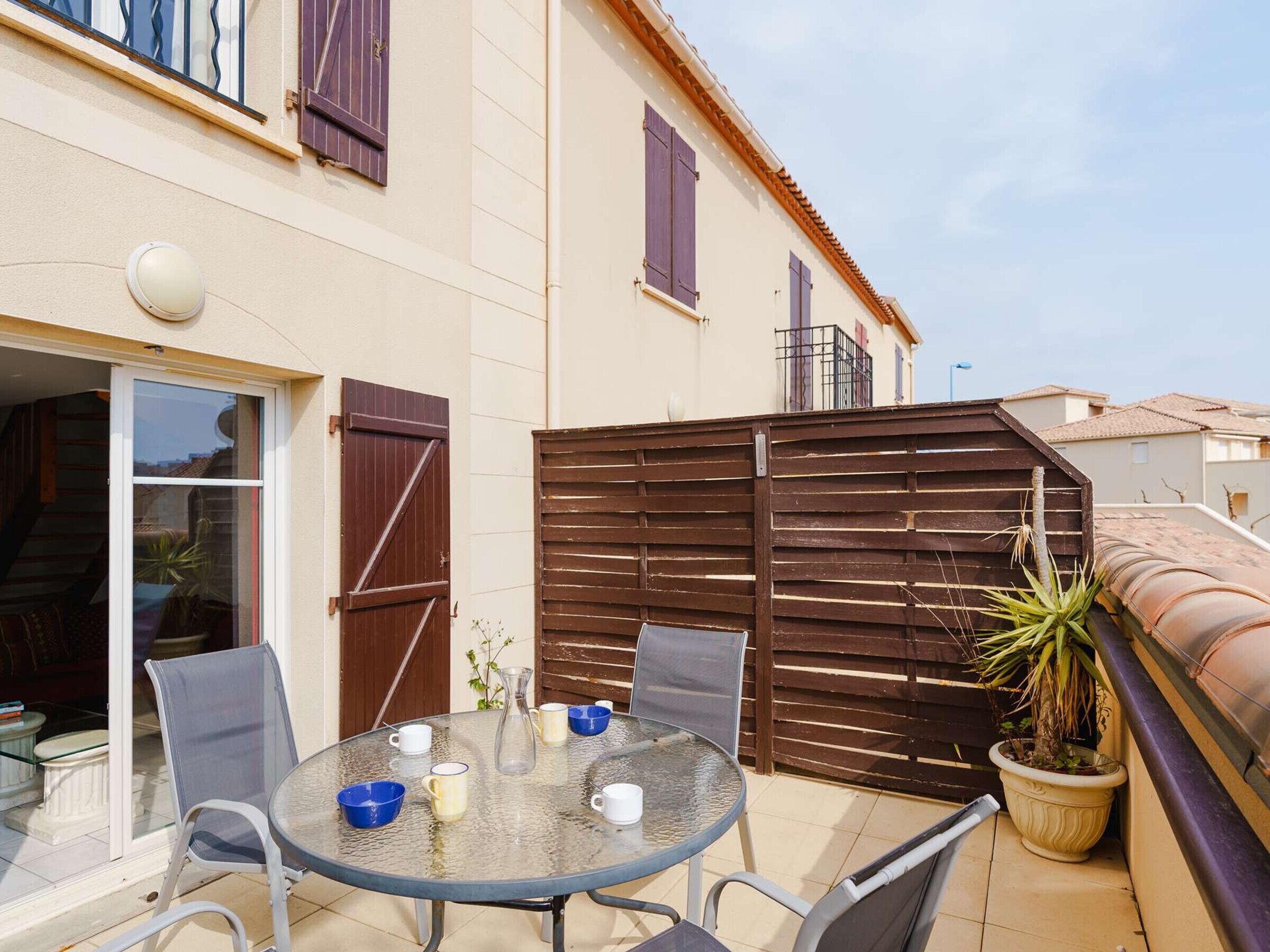Une table en verre, des chaises et une plante en pot sur un balcon agrémenté d’une clôture en bois.