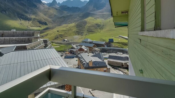 A view from a balcony overlooking a valley with mountains in the background, a small town below, and a building with green siding to the right.
