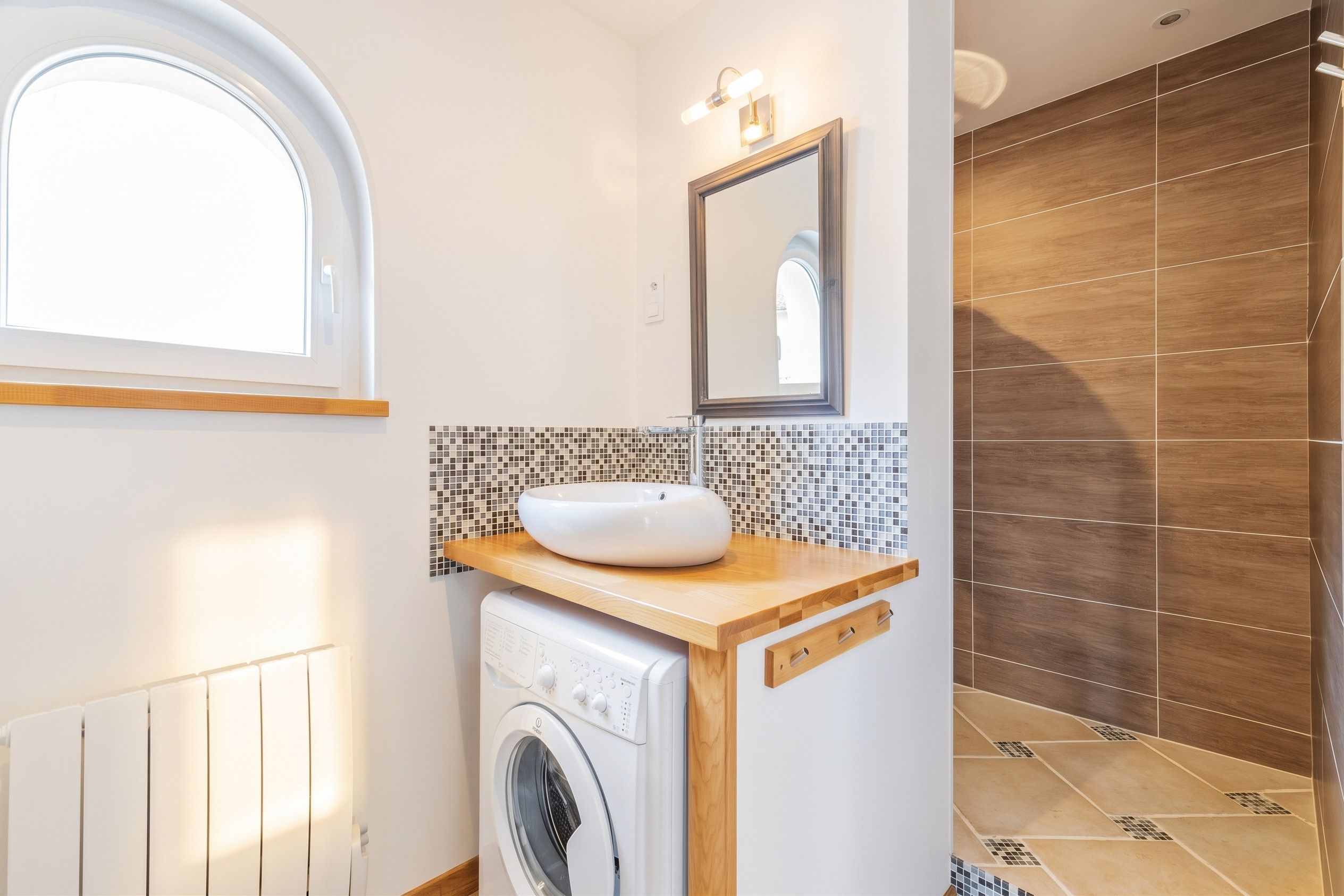 A compact bathroom with a built-in washing machine, a sink with a mosaic backsplash, and a mirror above it.