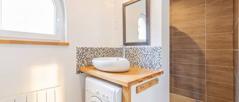 A compact bathroom with a built-in washing machine, a sink with a mosaic backsplash, and a mirror above it.