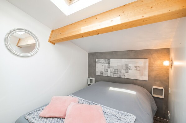 A bedroom with a skylight, a bed with pillows, a wall-mounted mirror, and a framed abstract artwork.