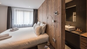A modern hotel room with a wooden panel wall, a large bed with white linens, a bedside table with a glass of water, a small bedside table with books, and a bathroom with a sink and mirror.