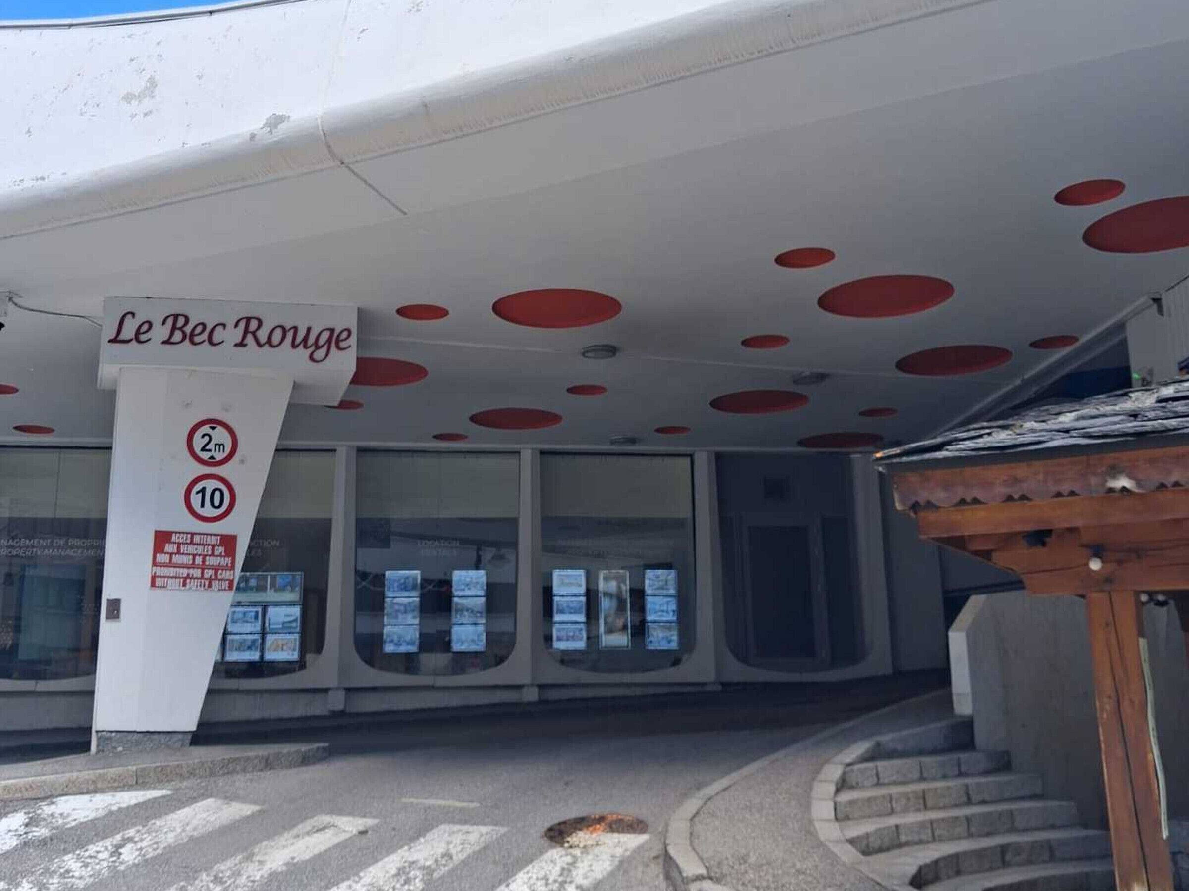 A building with a distinctive red and white color scheme, a sign reading 'Le Bec Rouge,' and a pedestrian crossing.