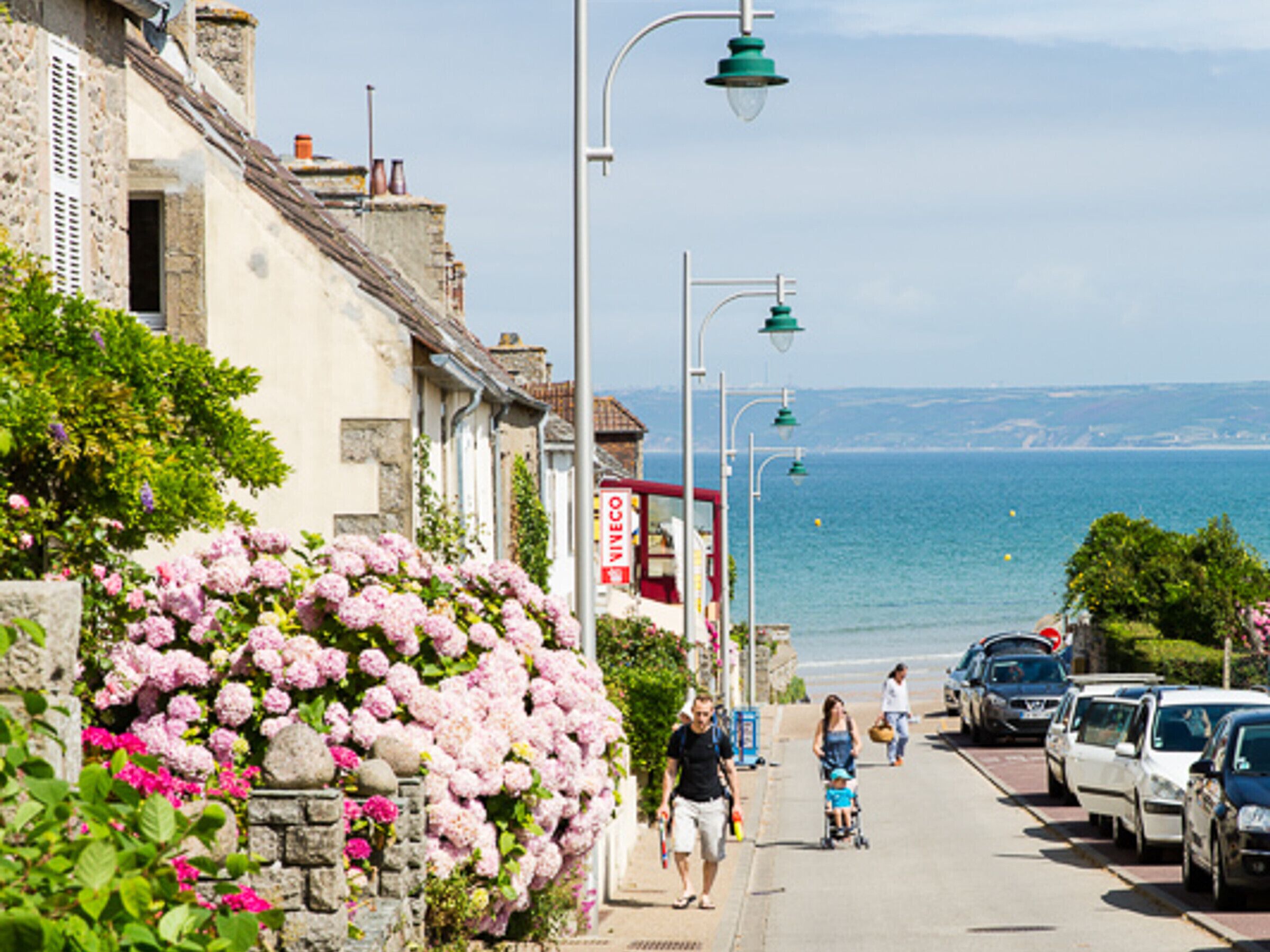 A coastal street with parked cars, pedestrians, and a row of buildings with stone facades and flowering plants.