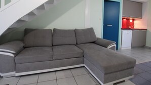 A modern living room with a grey sectional sofa, a staircase, and a small kitchenette with a red light. - Cozy cottage near the beach, wifi, pets allowed (Siouville-Hague)