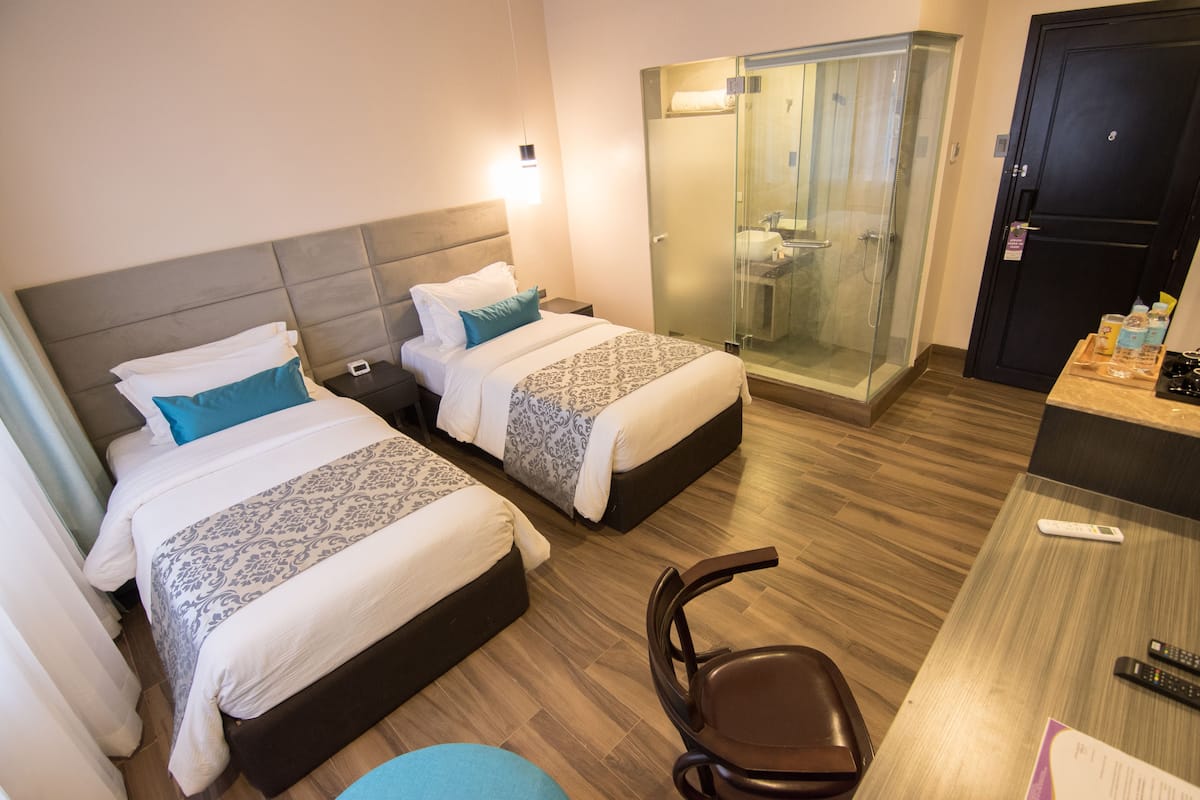 A hotel room with two beds, a shower area, a desk with a chair, and a remote control on the desk.