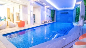 Une piscine intérieure moderne, avec une eau d’un bleu cristallin, entourée d’espaces de détente aux couleurs blanche et orange, et un mur de pierre agrémenté d’un jardin vertical verdoyant.
