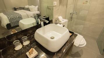 A modern bathroom with a large white sink, a glass shower enclosure, and a bed with white linens.