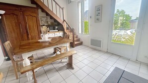 A dining area with a wooden table and benches, a kitchen in the background, and a view through a glass door. - Charming Villa with Garden, Steps to Beach, Near Town, Free WiFi (Jullouville)