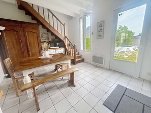 Un coin repas avec une table et des bancs en bois, une cuisine à l’arrière-plan et une vue à travers une porte vitrée.