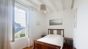 A bedroom with a wooden bed, a window with curtains, a bedside table, a lamp, and a painting on the wall. - Charming Villa with Garden, Steps to Beach, Near Town, Free WiFi (Jullouville)