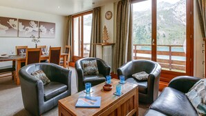 A spacious living area with leather sofas, a wooden coffee table, and a dining table set for four. Large windows offer a view of a mountainous landscape.