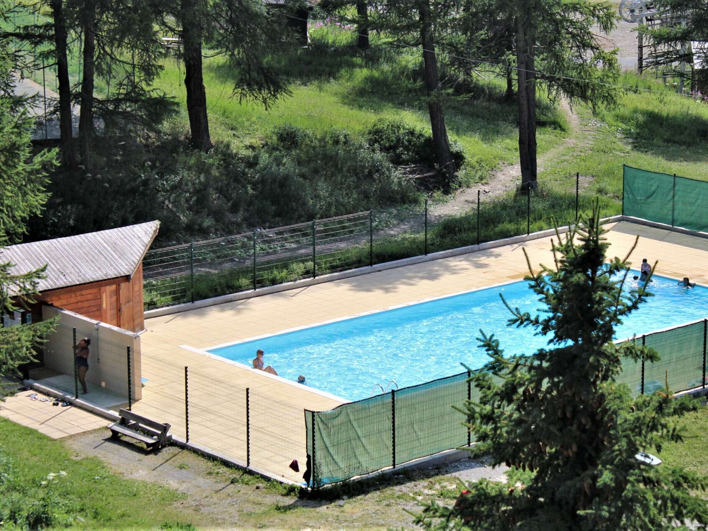 A rectangular swimming pool with a wooden deck and a small building with a sloped roof.