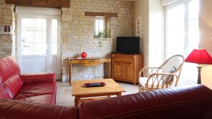 Un salon chaleureux avec un mur en pierre, une table basse en bois, un canapé en cuir rouge, un meuble en bois, un téléviseur et un fauteuil en osier.