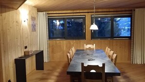 Un comedor con una mesa larga, sillas, un lámpara colgante y ventanas con cortinas.