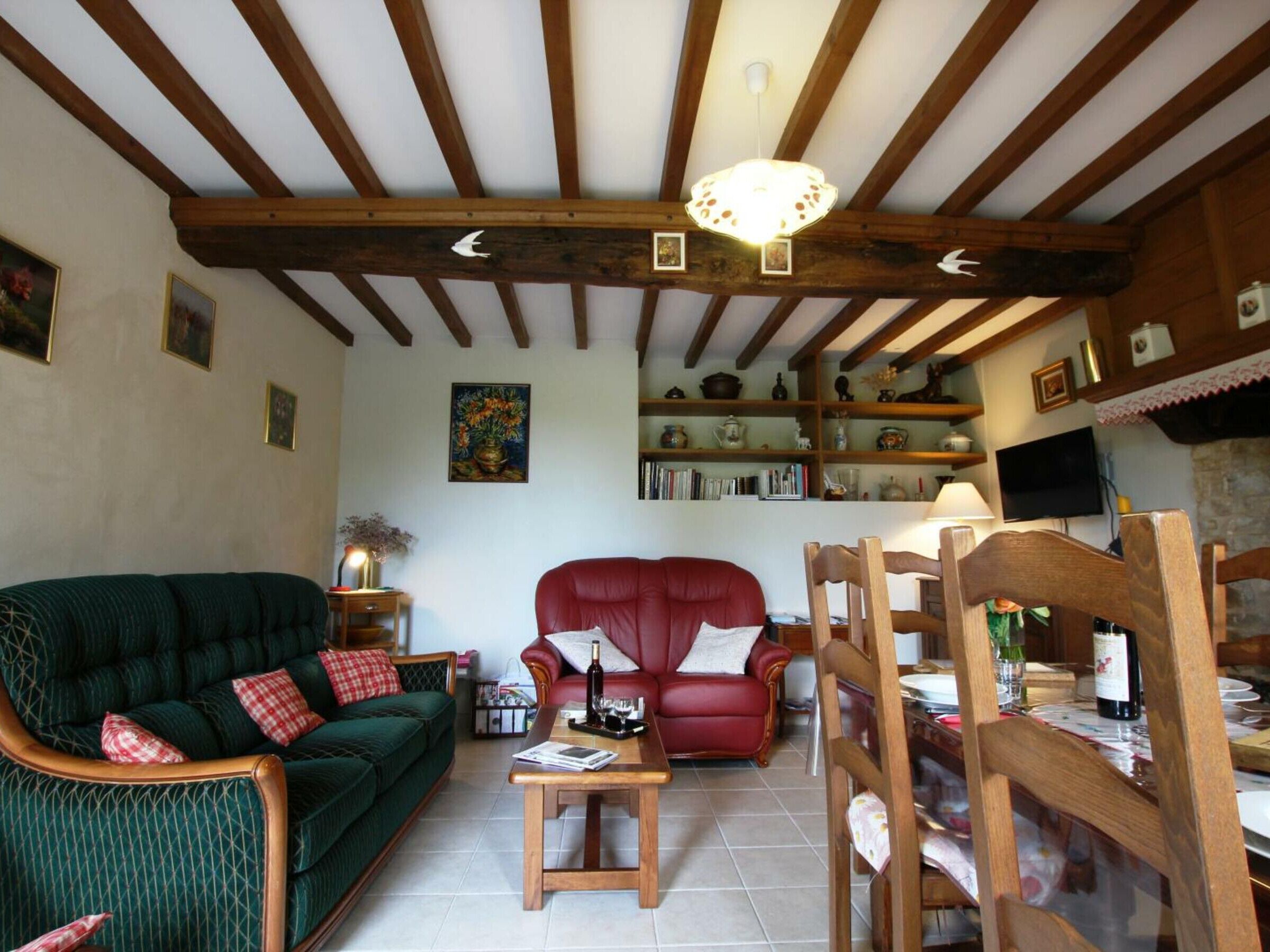 Uma sala de estar aconchegante com um sofá verde, uma poltrona vermelha e uma mesa de centro de madeira. Há estantes com diversos objetos e uma televisão na parede.