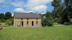 A two-story house with a stone facade, a grey roof, and a chimney. There is a small patio with a table and chairs, and a lawn in the foreground. - Quiet house with garden and parking near Lessay (Laulne)
