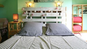 A bedroom with a bed, a bedside table with a lamp, a wooden headboard with decorative lights, a painting on the wall, and a wooden shelf with pillows.