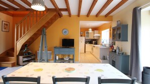 Un coin repas avec une table dressée pour quatre personnes, un escalier, une cuisine et un salon équipé d’un téléviseur.