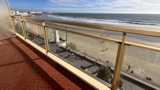 Een balkon met glazen hekwerk met uitzicht op een strand met een promenade en een klokkentoren.