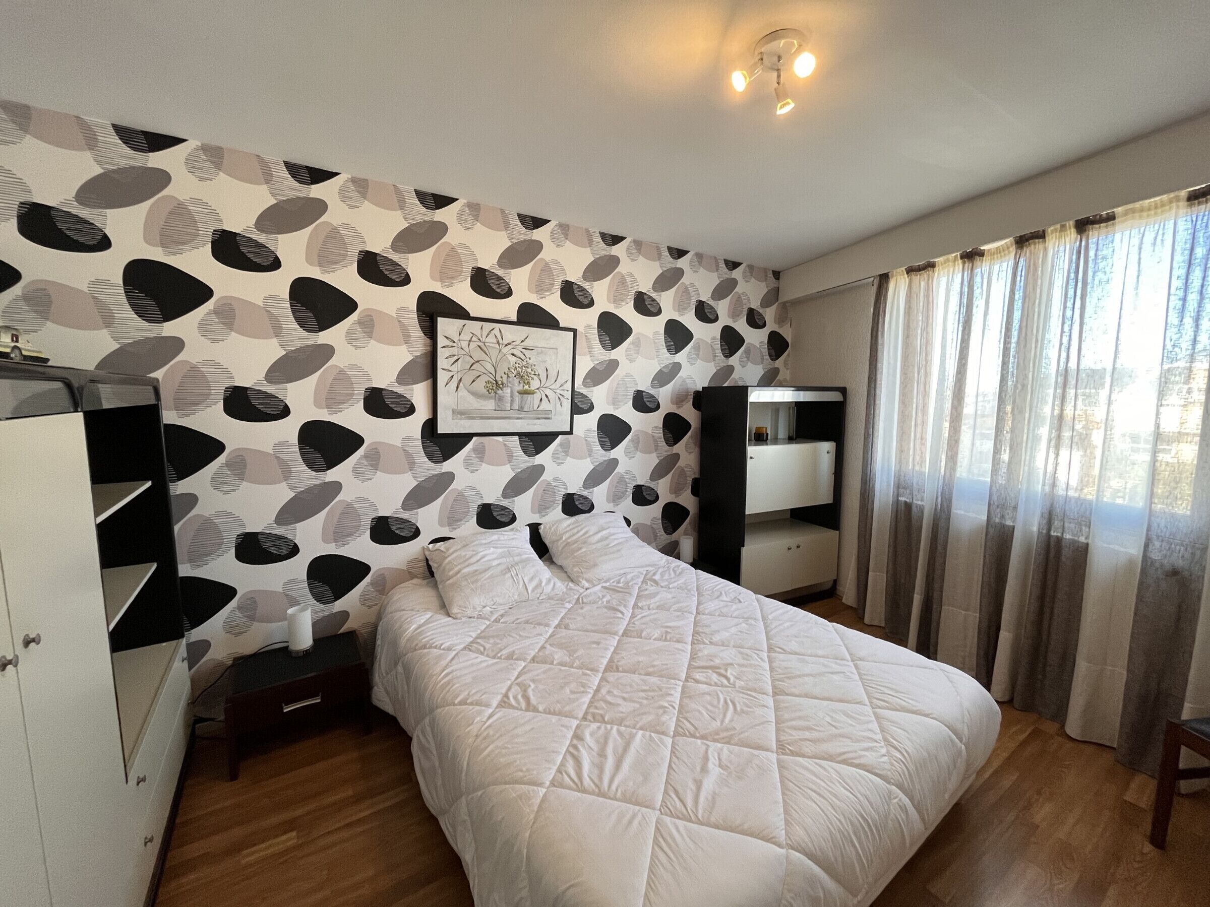 A bedroom with a bed, a nightstand, a wardrobe, a desk, and a window with curtains.