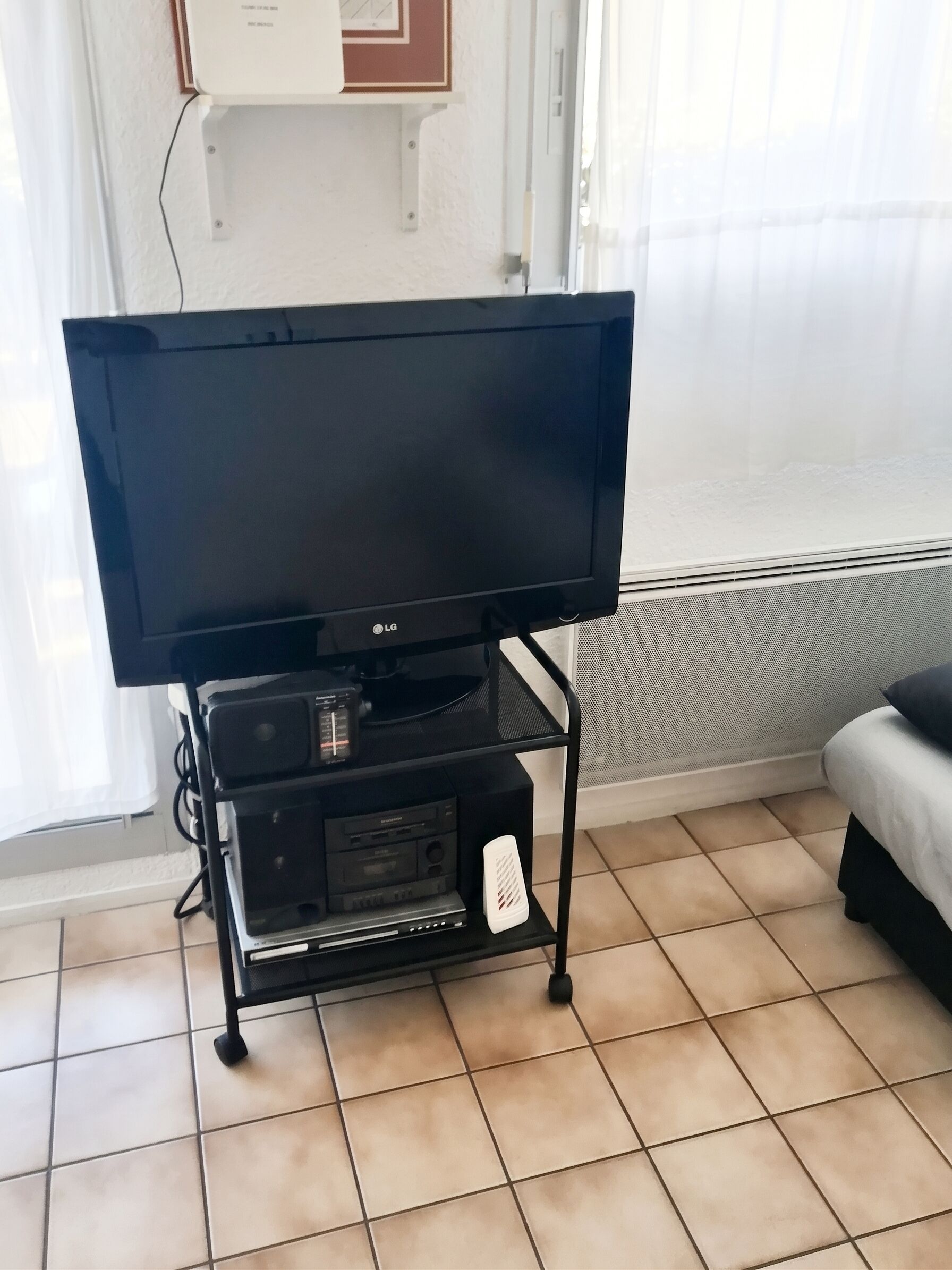 Un téléviseur posé sur un meuble, un lit, un sol carrelé et une fenêtre avec des rideaux.