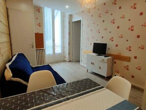Une pièce comprenant un lit, un téléviseur posé sur un meuble, une table à manger et une fenêtre avec des stores.
