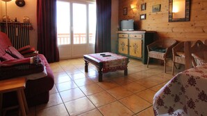 Una habitación acogedora con techo de madera, un sofá, una mesita, sillas de mimbre, un mueble con televisor y vistas al balcón.