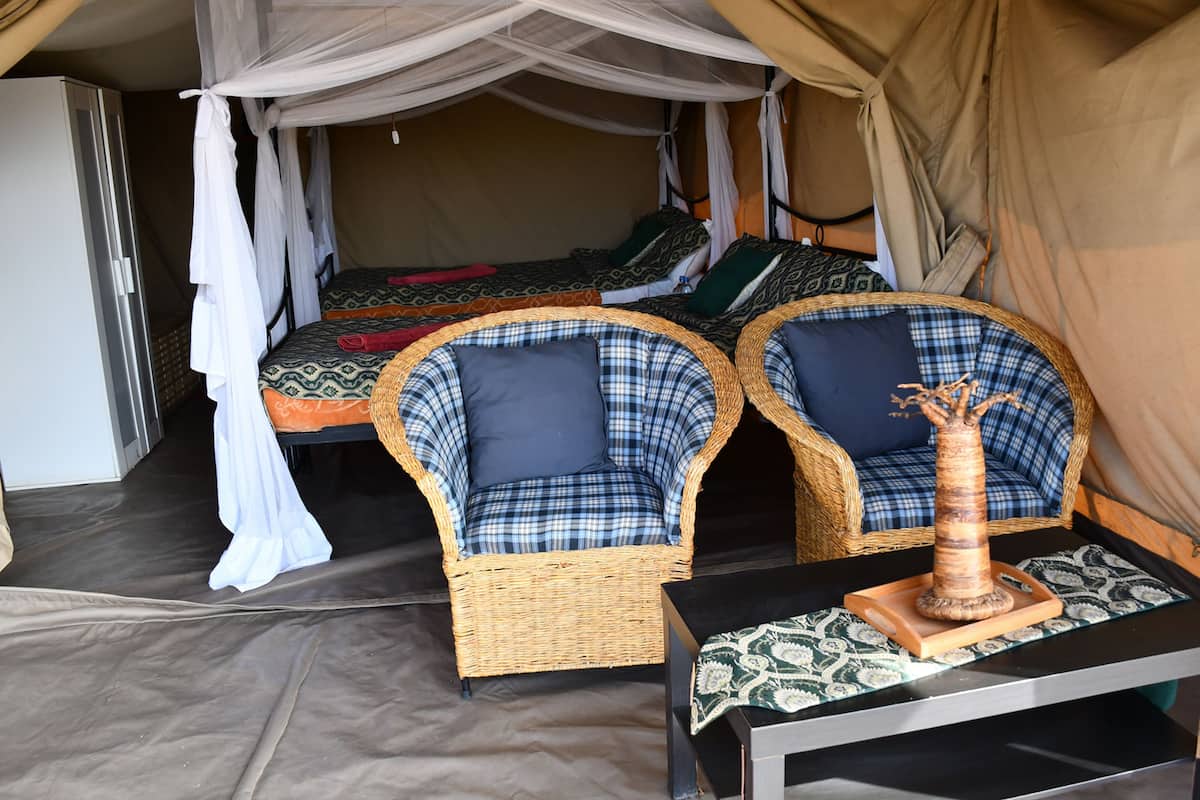 A room with two beds, each with patterned bedding, a wicker chair with a cushion, and a small table with a decorative item.