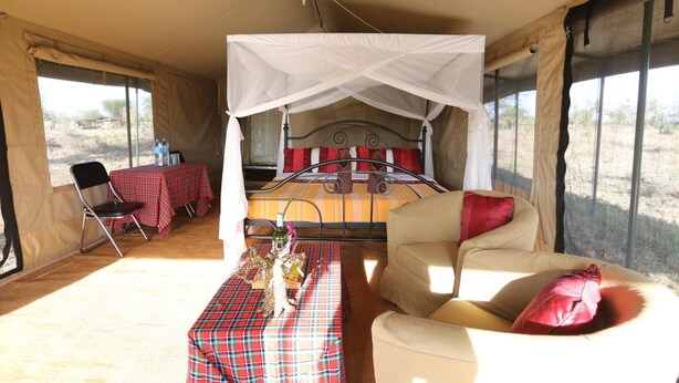 A tented room with a bed, a table with a red and black checkered tablecloth, a chair, and a sofa with red cushions. - Serengeti Wildebeest Camp (Serengeti)