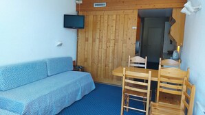 Una stanza piccola con soffitto in legno, una TV a parete, un divano blu, un tavolo in legno con sedie e un tappeto blu.