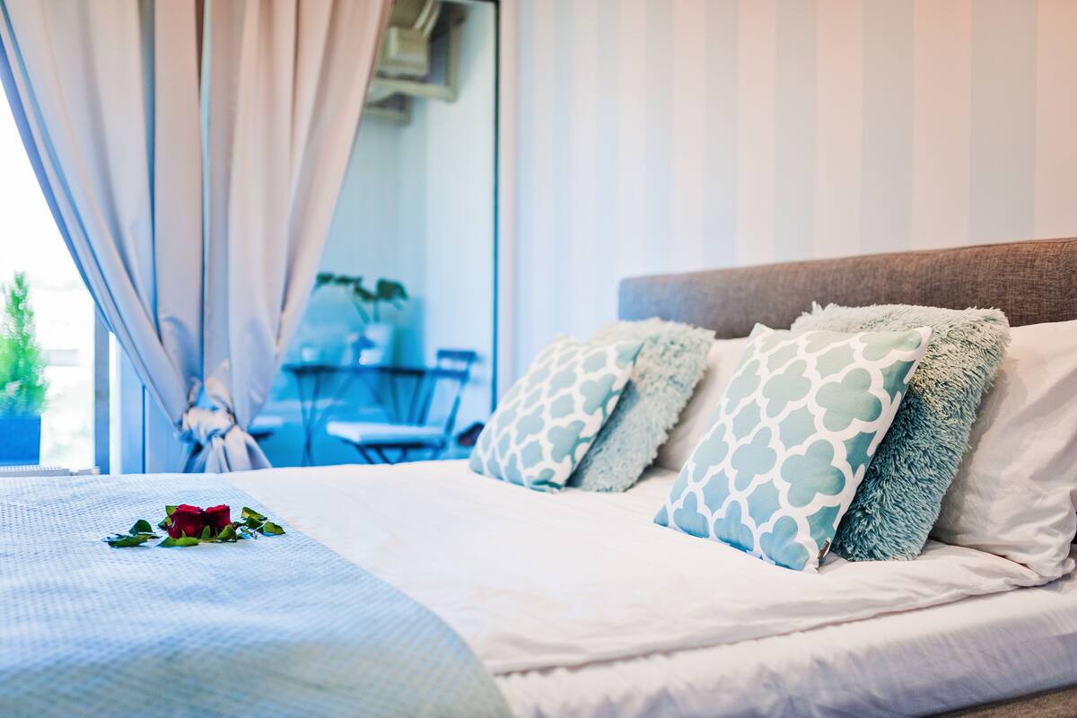A neatly made bed with patterned pillows and a single rose, a window with curtains, and a view of a balcony area with a table and chairs.
