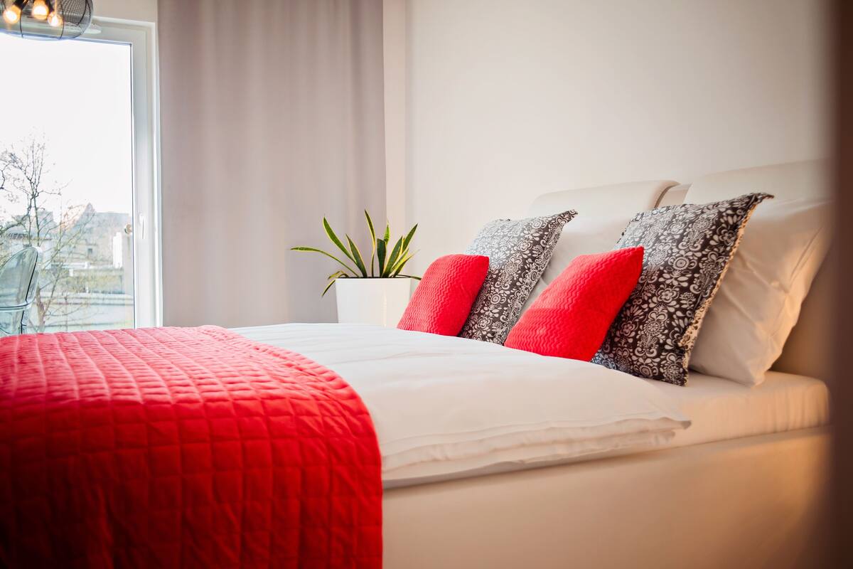 A neatly made bed with a red blanket and patterned pillows, a potted plant, and a window with a view of buildings.