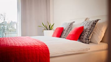A neatly made bed with a red blanket and patterned pillows, a potted plant, and a window with a view of buildings.