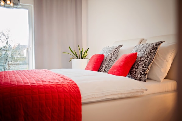 A neatly made bed with a red blanket and patterned pillows, a potted plant, and a window with a view of buildings.
