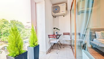 A balcony with potted plants, a table with chairs, a window with a view of greenery, and an air conditioning unit.